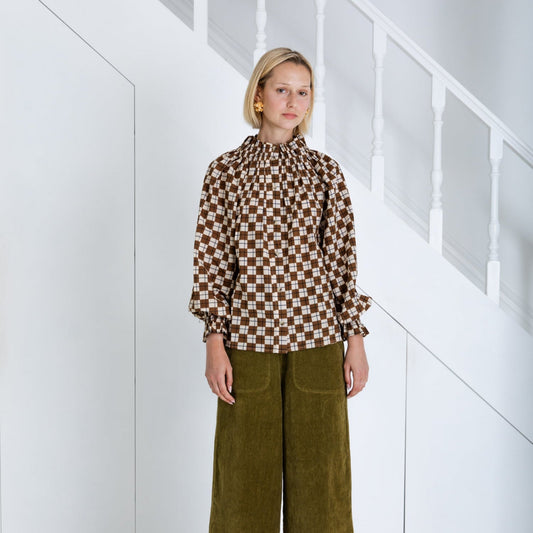 Woman wearing a brown checkered blouse and green pants against a white geometric background