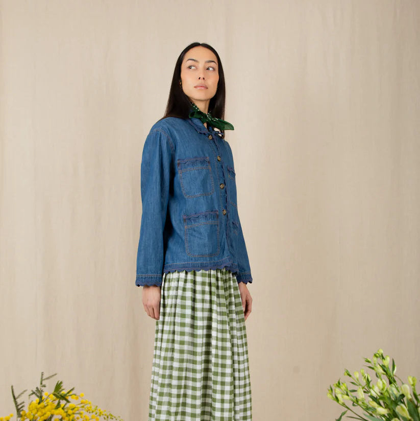 Woman wearing a blue denim jacket and green checkered dress standing against a beige wall with floral decorations.