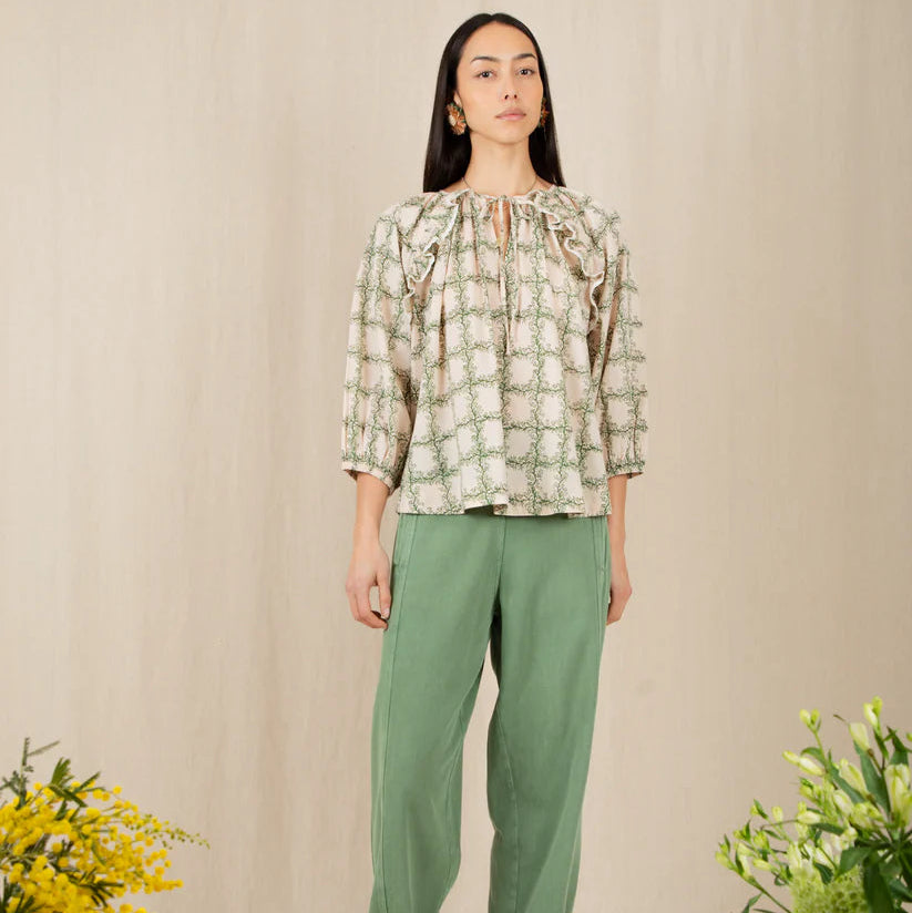 Woman wearing a patterned blouse and green pants standing in a room with flowers.