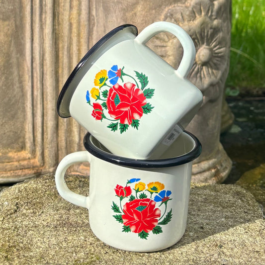 Two white enamel mugs with floral designs stacked on a sandy surface.
