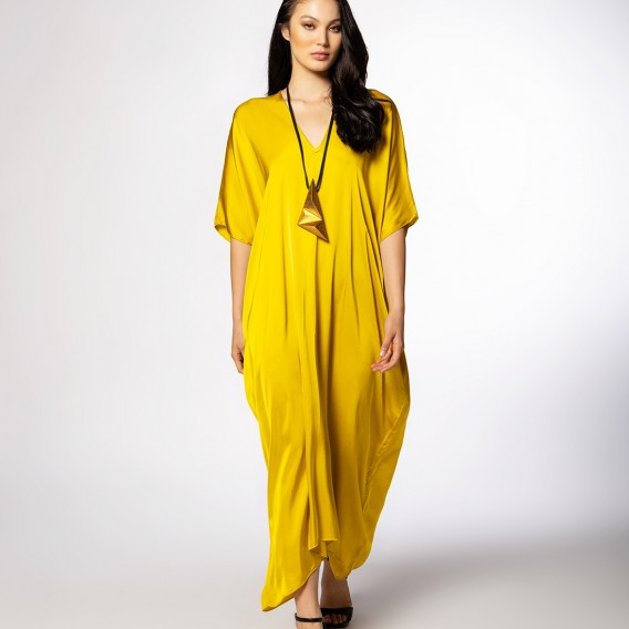 A woman modeling a bright yellow kaftan dress with short wide sleeves and a V-neckline, paired with a long black pendant necklace and black strappy heels, against a light gray studio background.