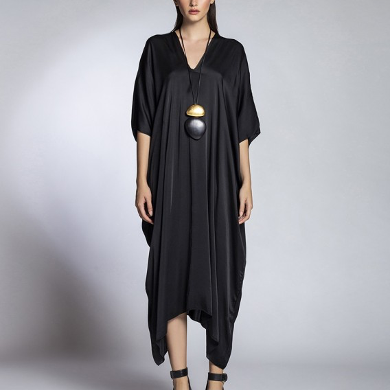 A woman modeling a black draped kaftan dress with a V-neckline and wide short sleeves, styled with a long pendant necklace and black strappy high-heeled sandals against a light gray studio background.
