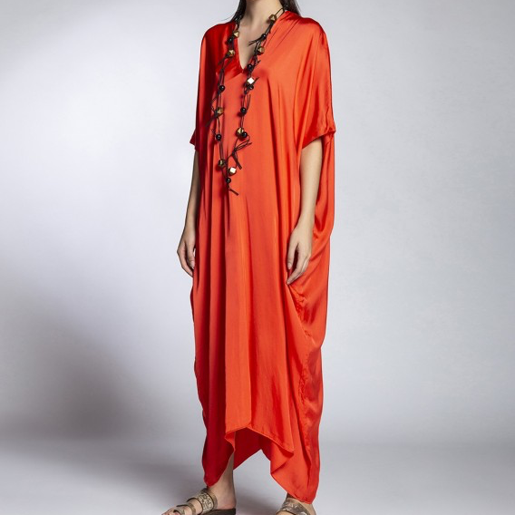 A woman modeling a flowing, draped orange-red maxi dress with short wide sleeves and a loose, draped silhouette, styled with a long beaded necklace and sandals against a light gray studio background.