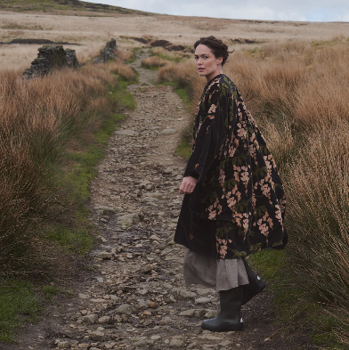 A woman walks along a rocky path through a grassy field, wearing a long black coat with a pink floral pattern, a gray skirt, and dark rubber boots.