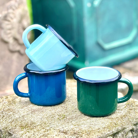 Three enamel mugs in blue, green, and white on a stone surface with a blurred background.
