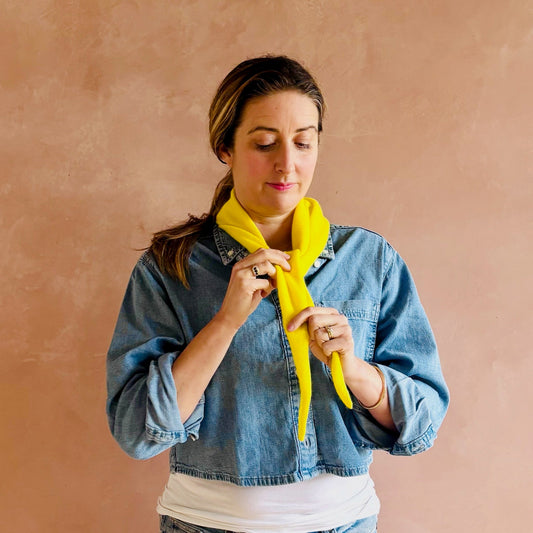 Woman wearing a denim jacket and white shirt, holding a yellow scarf against a beige background