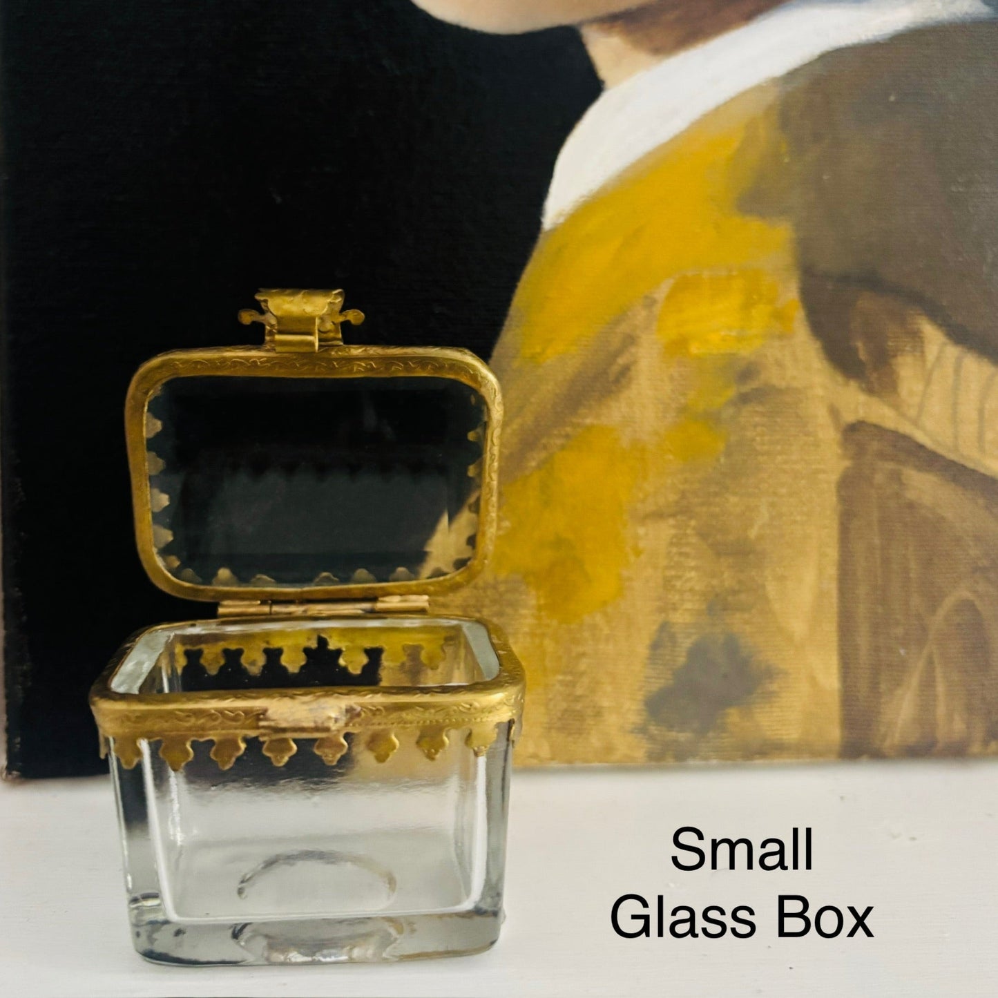 Small glass box with gold lid on a white surface with a blurred painting in the background