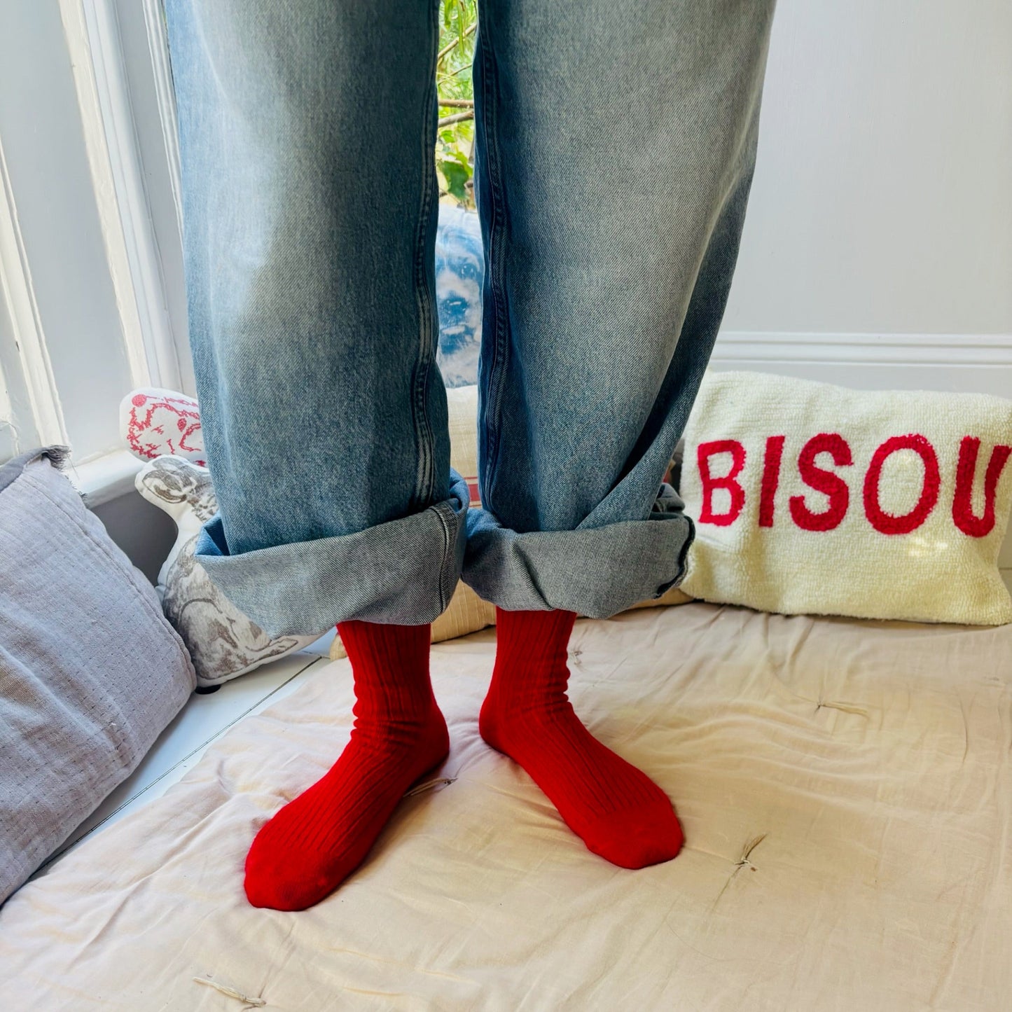 Person wearing red socks and blue jeans standing on a bed with a pillow that says 'BISOU'.