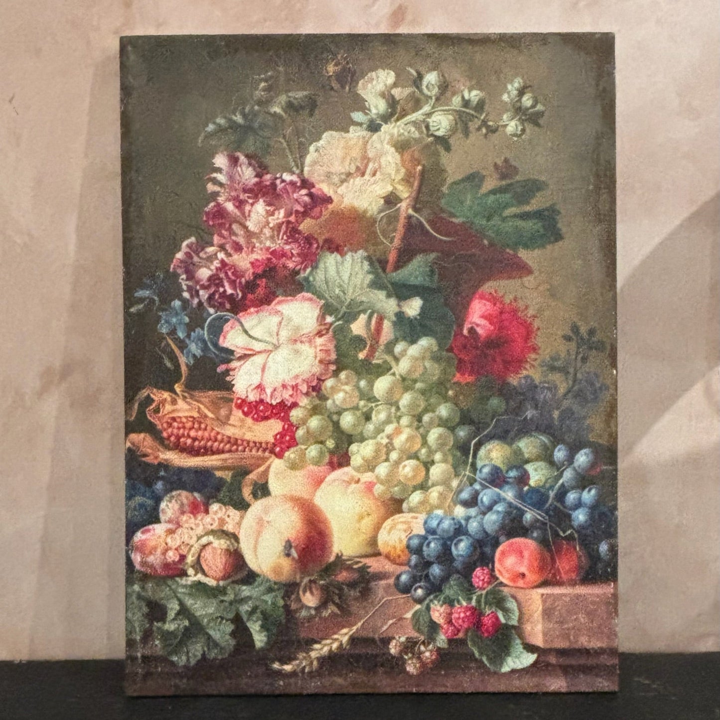 Framed still life painting of fruits and flowers on a textured wall.