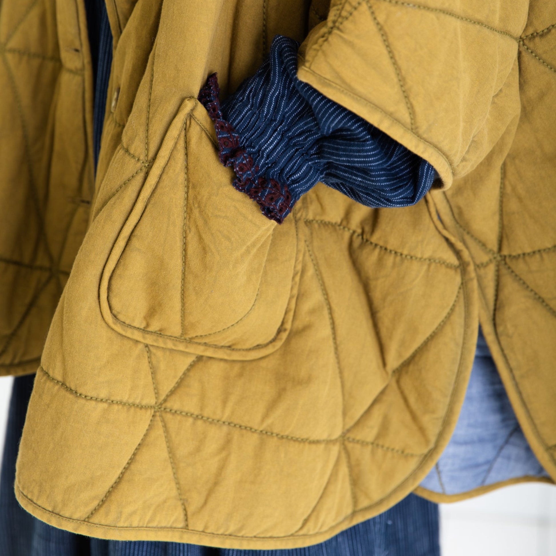 Close-up of a mustard yellow quilted jacket with a blue fabric inside the pocket on a white background
