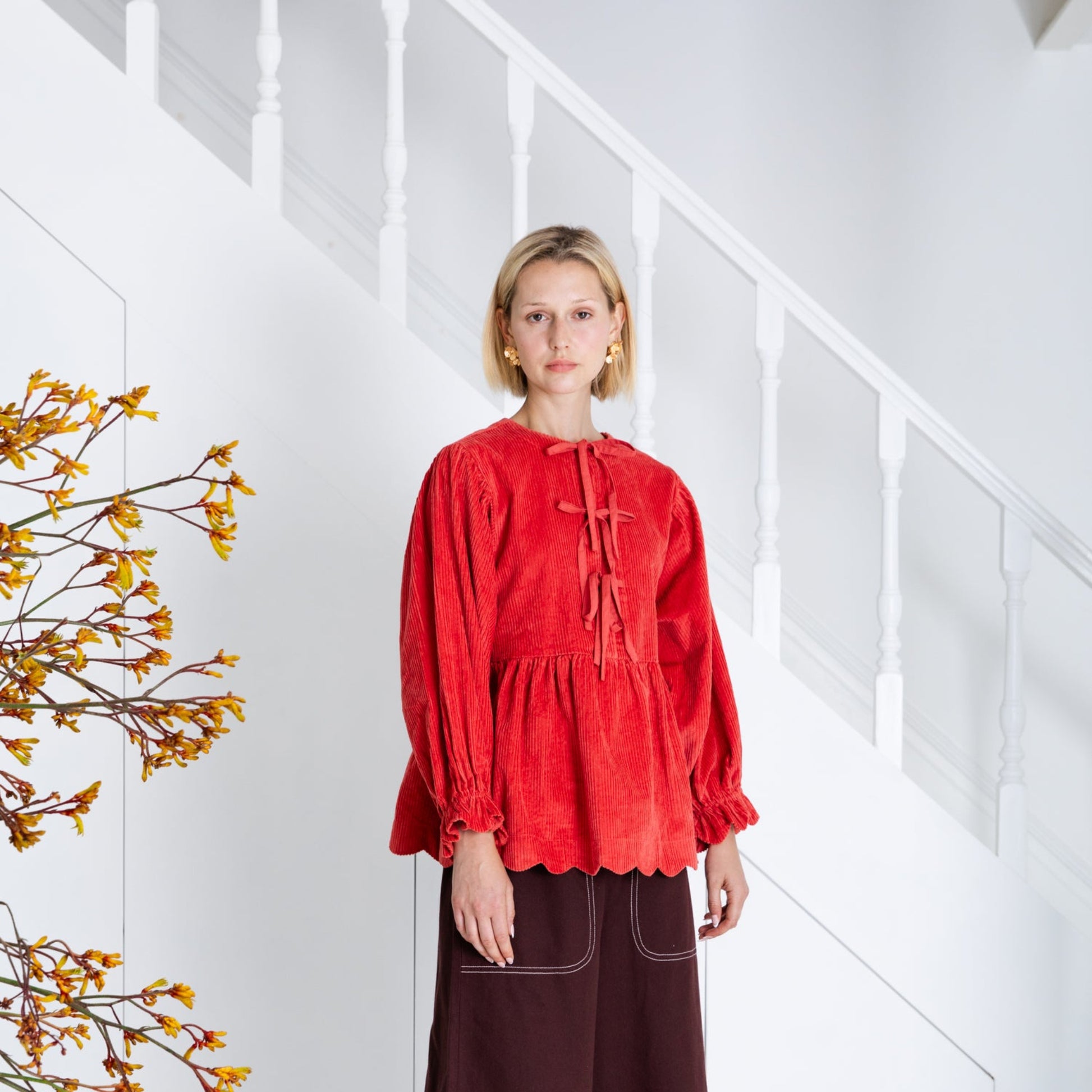 Woman wearing a red top and brown skirt standing in a minimalistic room with white walls and wooden floor.
