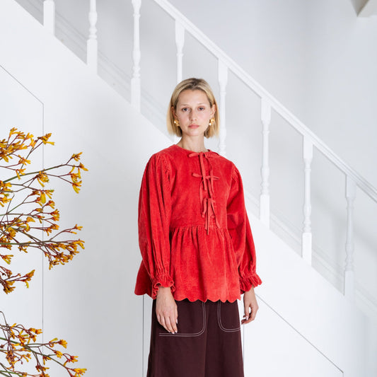 Woman wearing a red top and brown skirt standing in a minimalistic room with white walls and wooden floor.