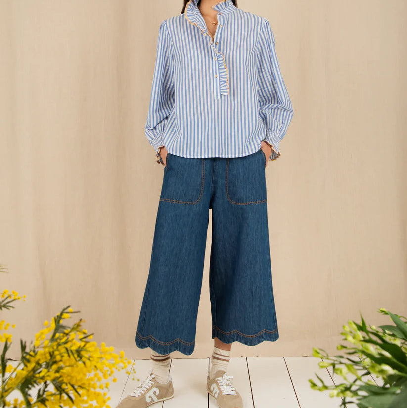 Woman wearing a blue and white striped shirt and blue jeans standing against a beige wall with yellow flowers on a wooden floor.