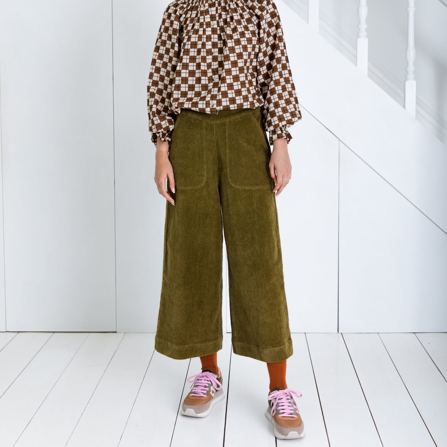 Person wearing a patterned shirt and green pants standing in a minimal white room.