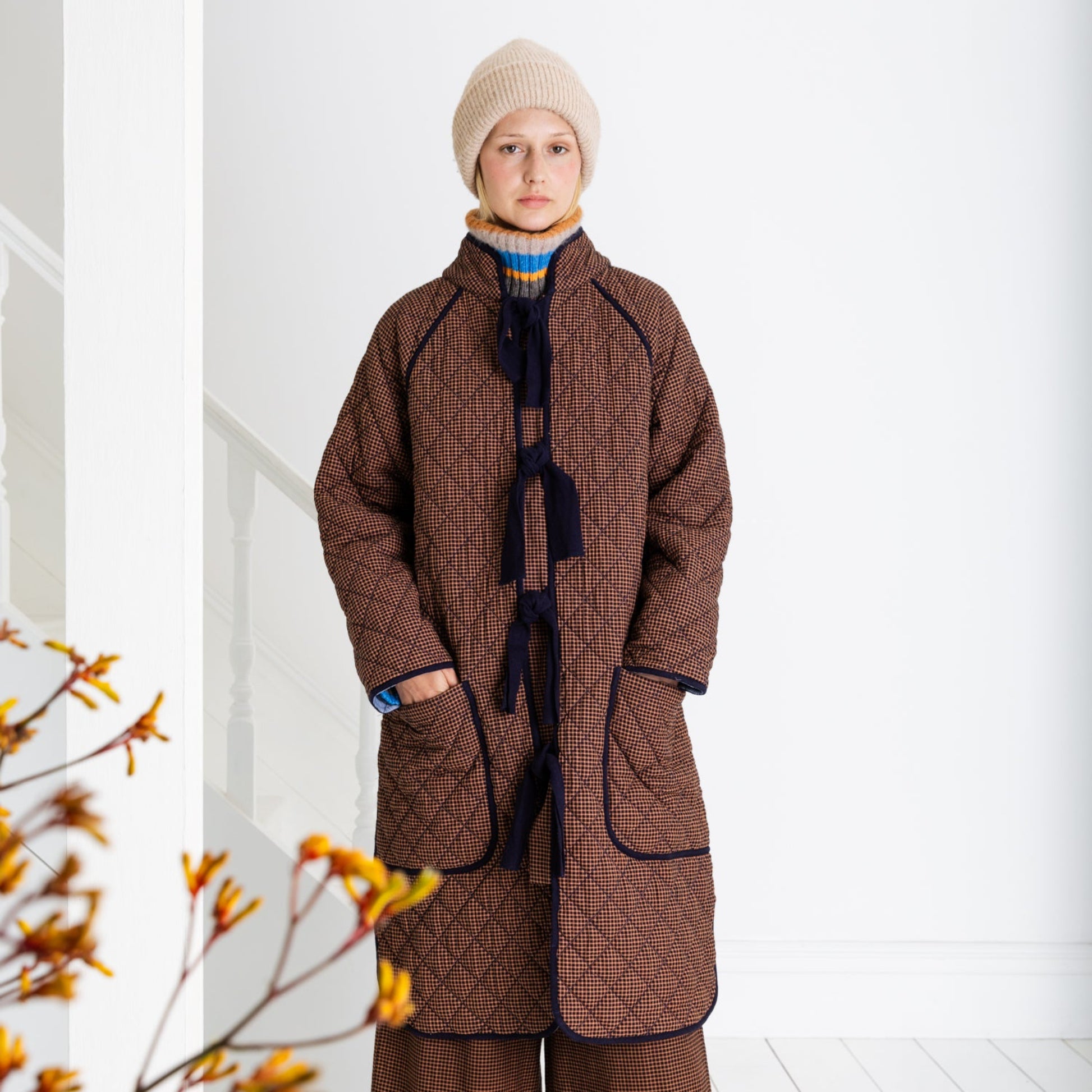 Person wearing a long brown coat and hat in a white room with a branch of yellow flowers.