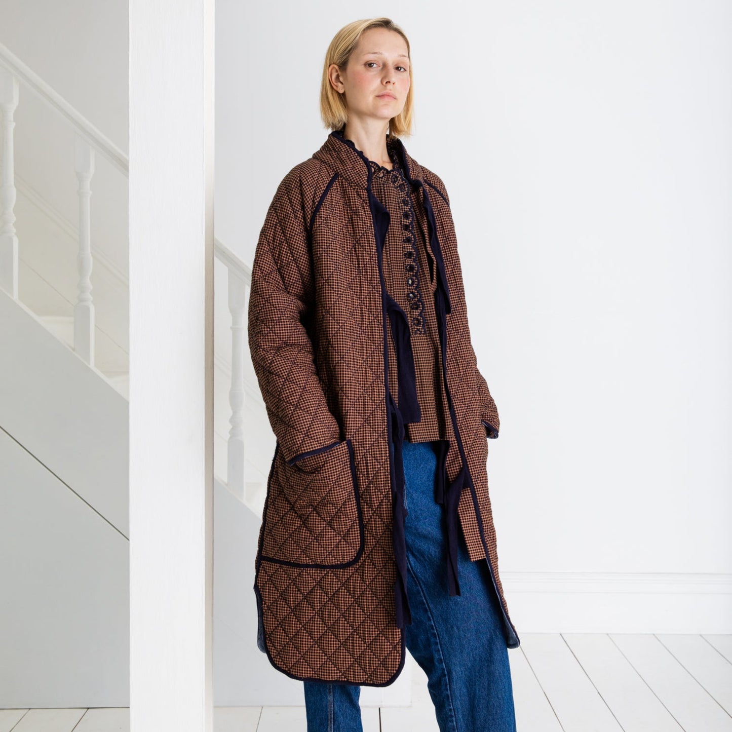 Person wearing a long brown quilted coat in a minimal white room