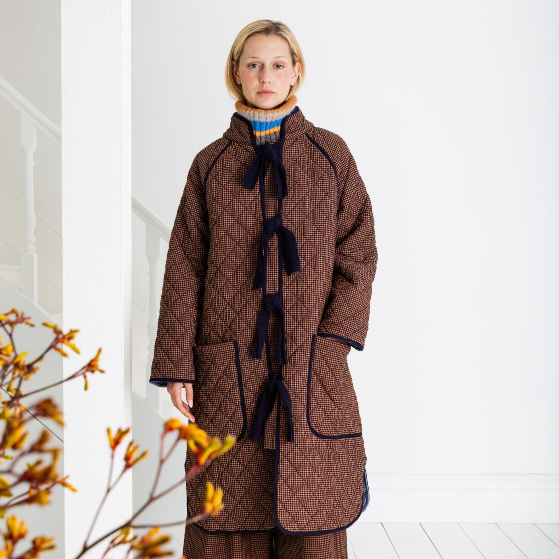 Woman wearing a brown quilted coat in a minimal white room with a branch of yellow flowers.