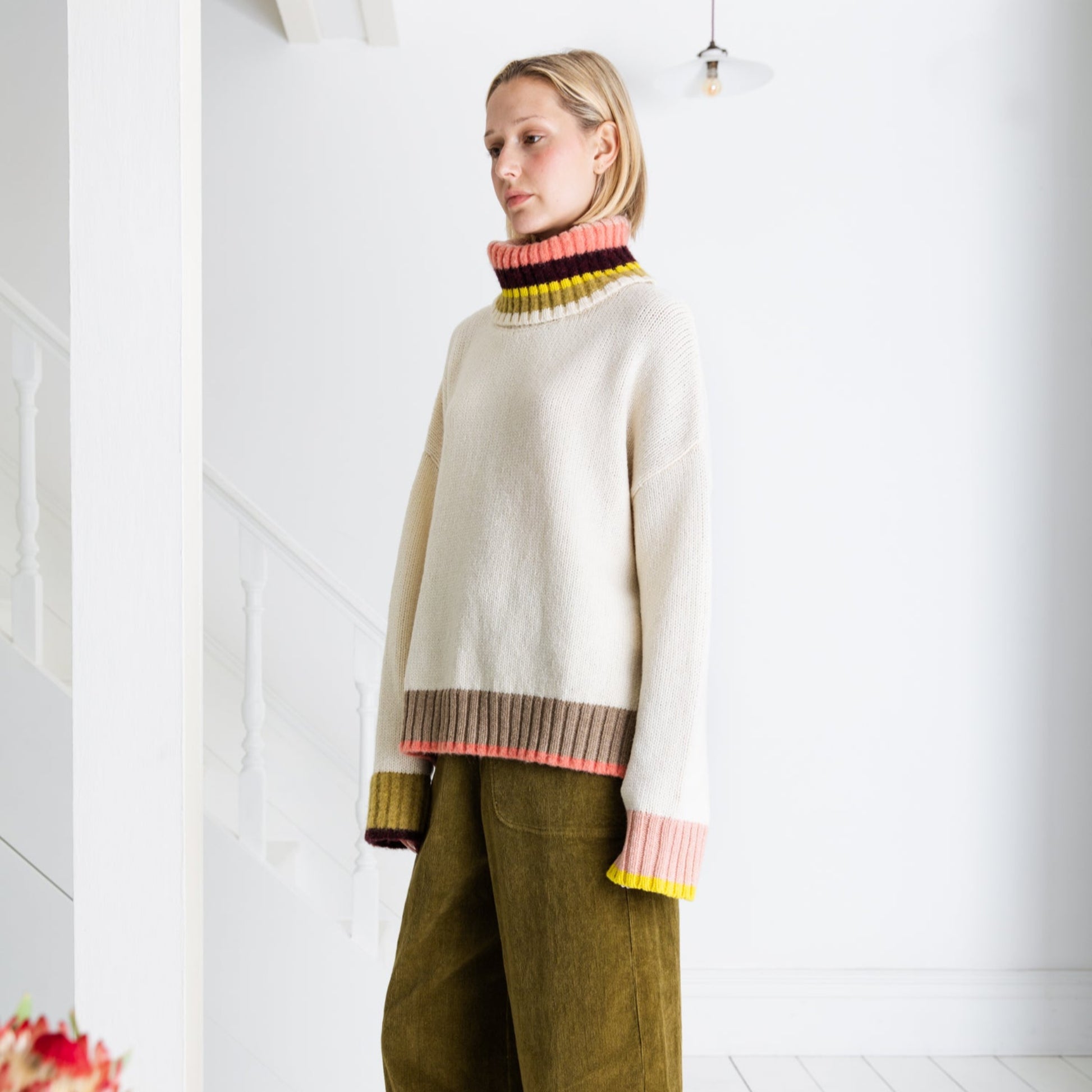 Woman wearing a colorful sweater and green pants in a minimal white room.