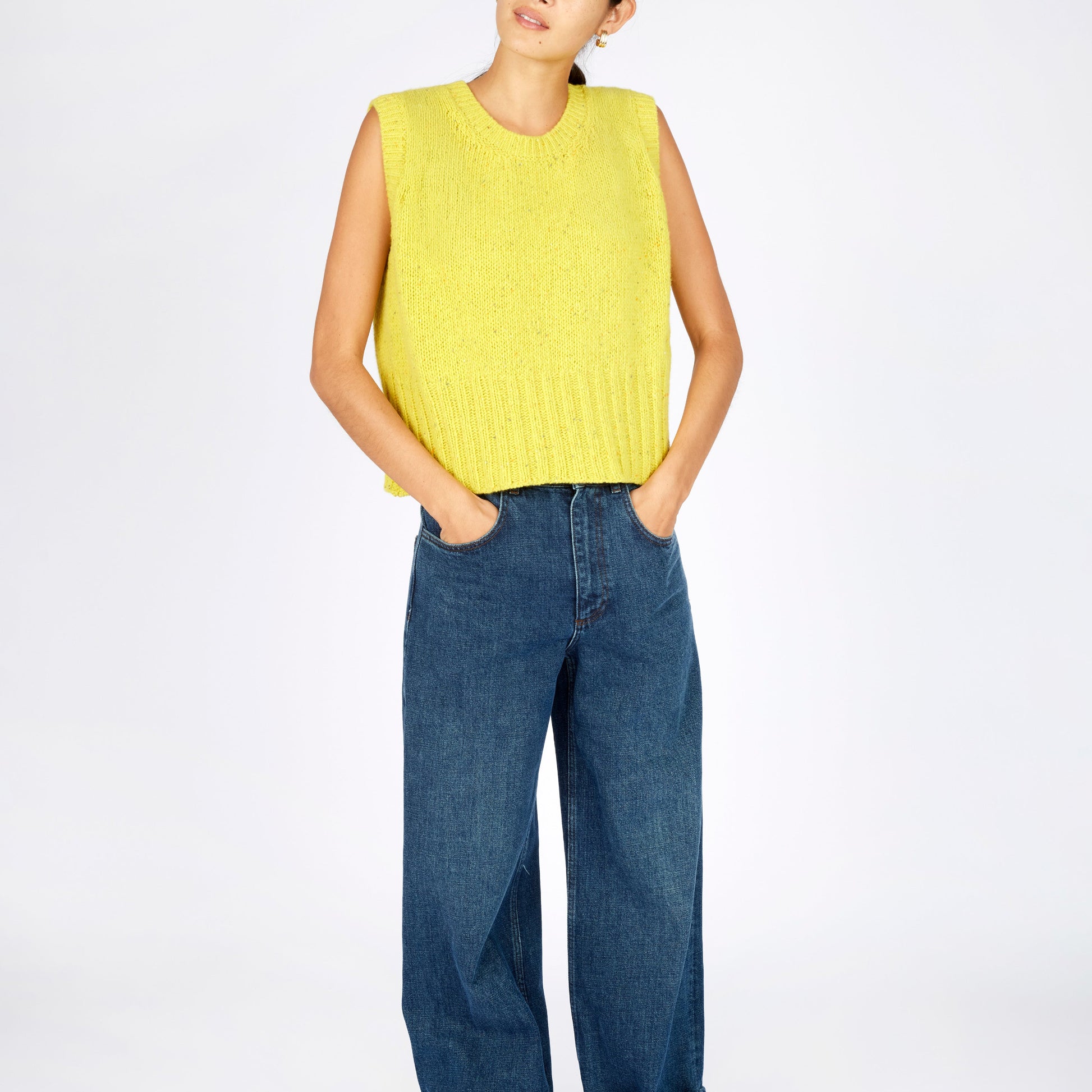 Person wearing a yellow sleeveless sweater and blue jeans on a white background