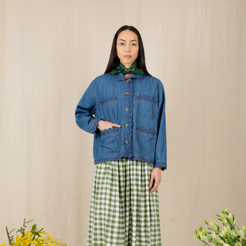 Woman wearing a blue denim jacket and green checkered pants standing against a beige wall with floral decorations.