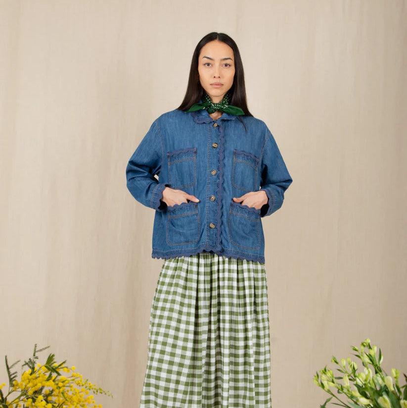 Woman wearing a blue denim jacket and green checkered dress standing against a beige wall with floral decorations.