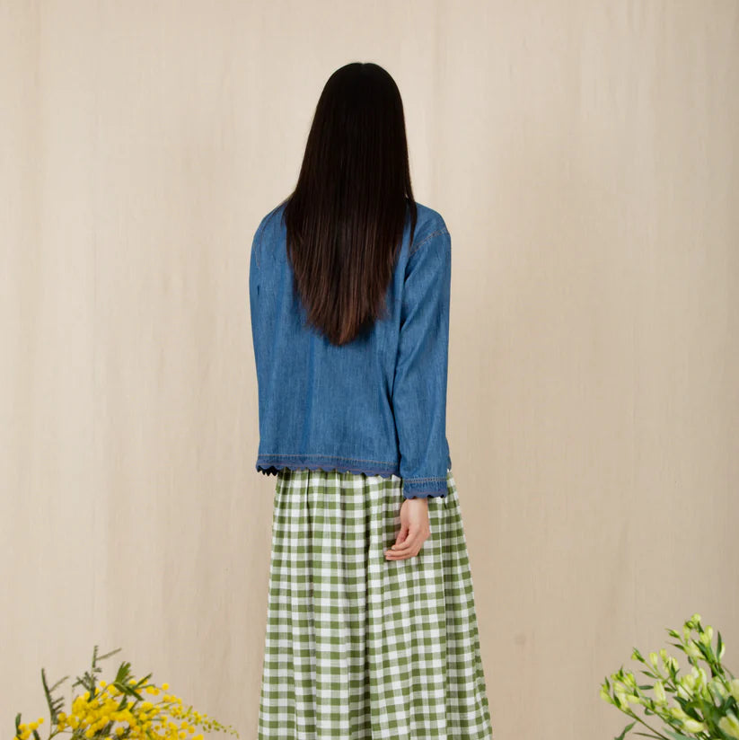 Person wearing a blue top and green checkered skirt standing in front of a beige wall with flowers on either side.