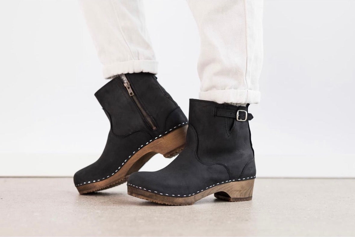 A pair of black suede ankle boots with brown wooden heels and white stitching, shown on a model wearing white pants against a clean white background.