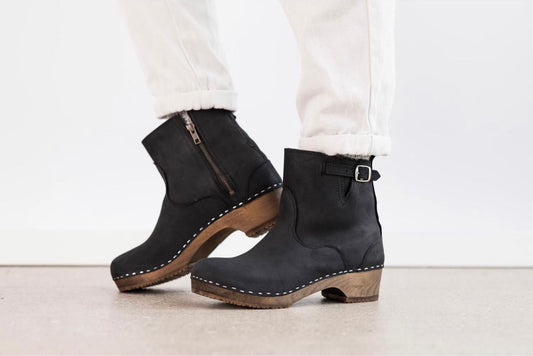 A pair of black suede ankle boots with brown wooden heels and white stitching, shown on a model wearing white pants against a clean white background.