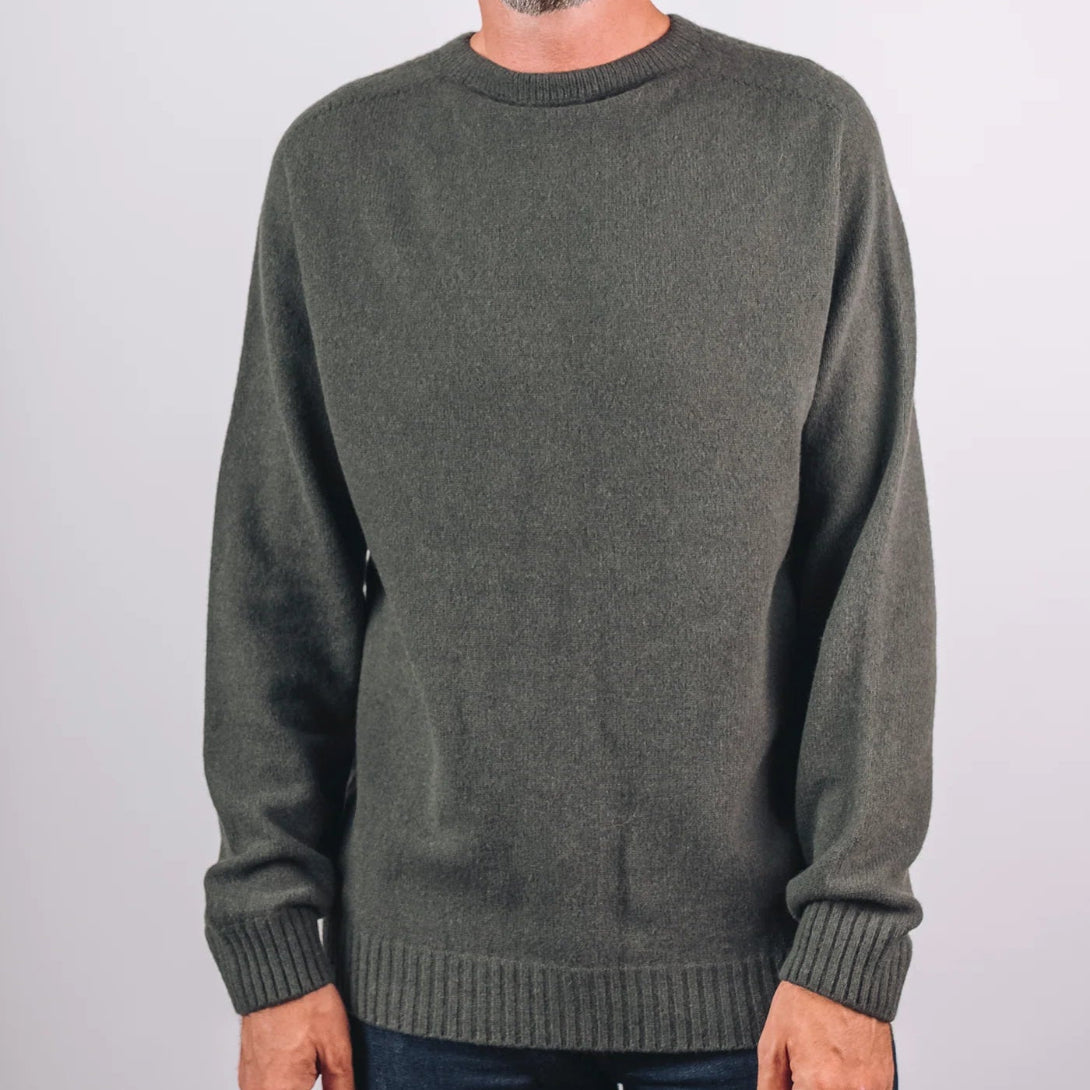 A man wearing a dark olive green knit crewneck sweater, a tan ribbed beanie, and dark blue jeans, standing against a light gray studio background.