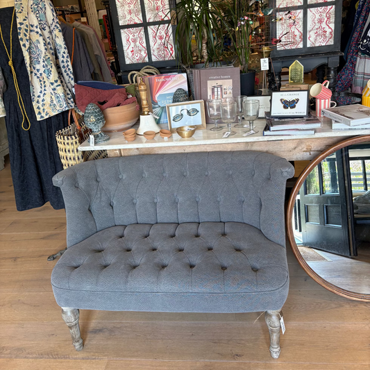 Gray tufted bench in a store setting with clothing and decor items.