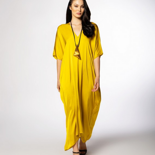 A woman modeling a bright yellow kaftan dress with short wide sleeves and a V-neckline, paired with a long black pendant necklace and black strappy heels, against a light gray studio background.