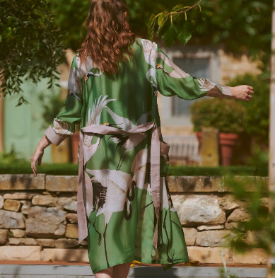 Person wearing a green patterned dress standing outdoors with trees and stone wall in the background