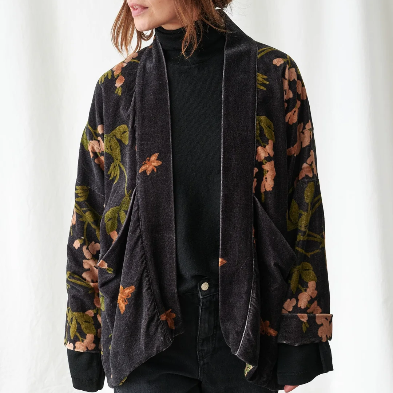 A woman wearing a dark charcoal velvet jacket with a muted floral pattern in green, orange, and pink, styled over a black turtleneck and dark jeans against a white curtain backdrop.