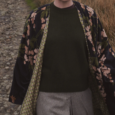 A woman standing outdoors on a path wearing a dark olive green knit sweater under a black floral kimono-style jacket with green patterned lining, paired with gray pleated trousers.