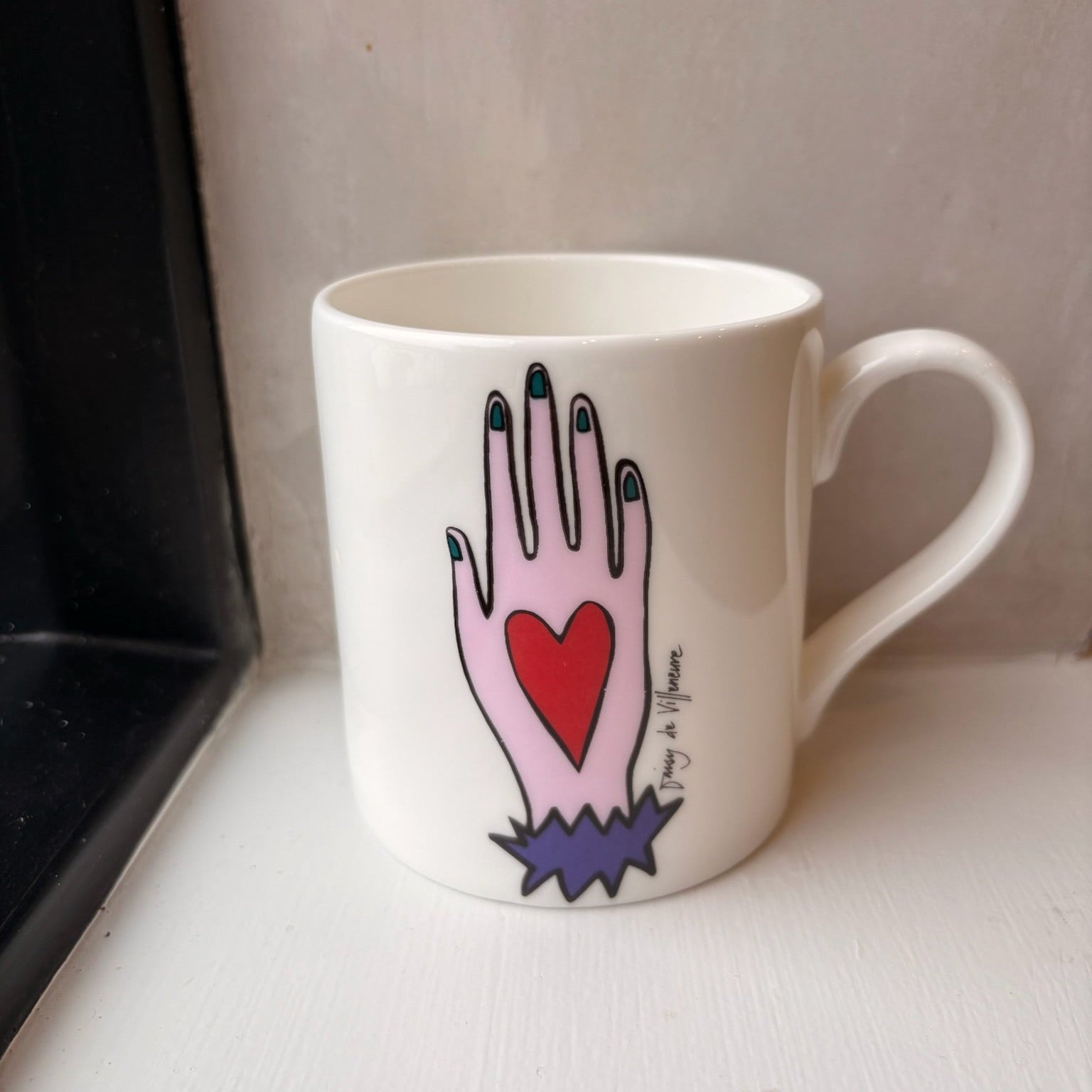White mug with a hand-drawn design of a hand holding a red heart on a white surface.