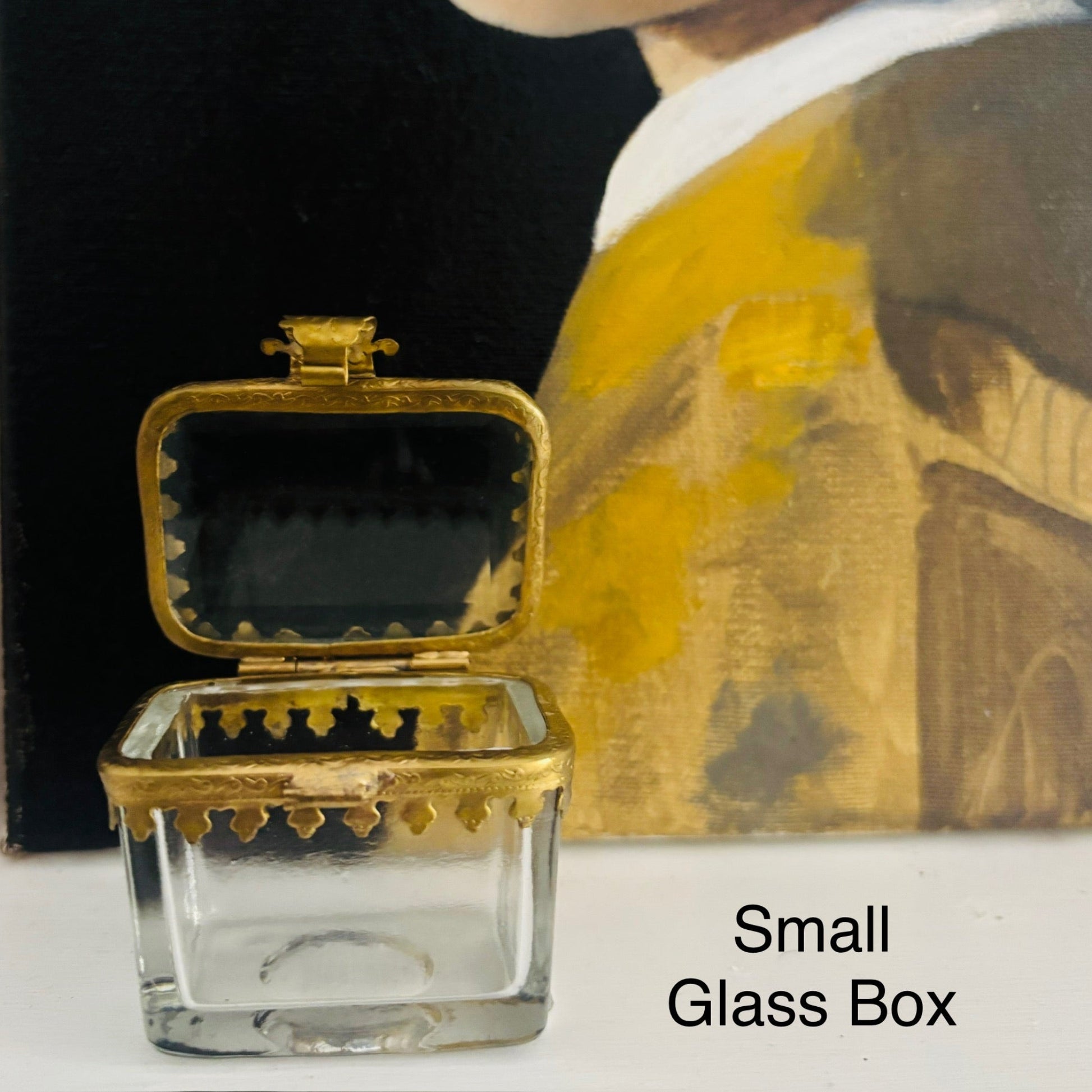 Small glass box with gold lid on a white surface with a blurred painting in the background