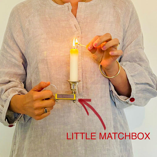 Person lighting a candle with a matchbox labeled 'LITTLE MATCHBOX' on a plain background