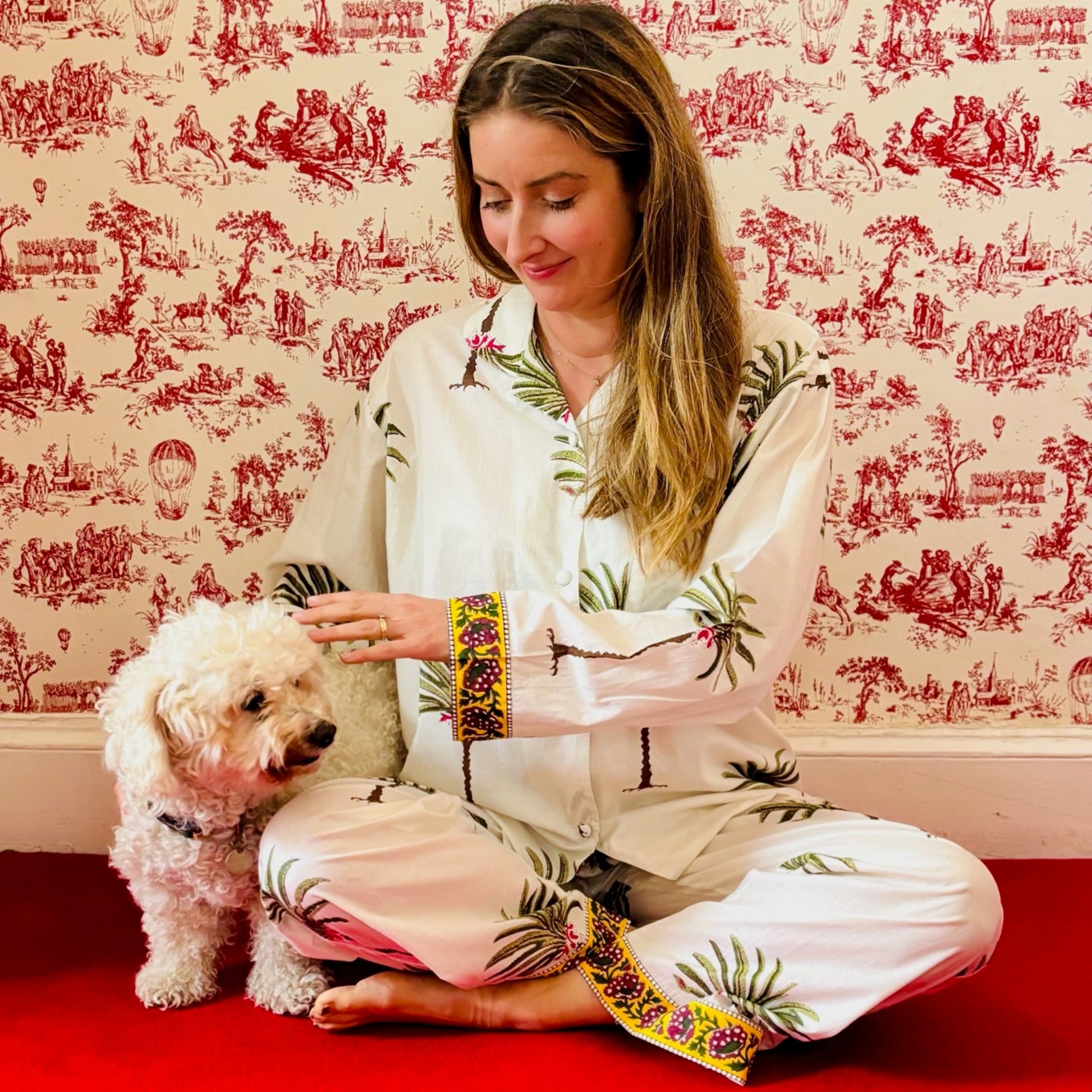 Woman in a floral robe petting a small dog against a decorative red and white wallpapered wall.
