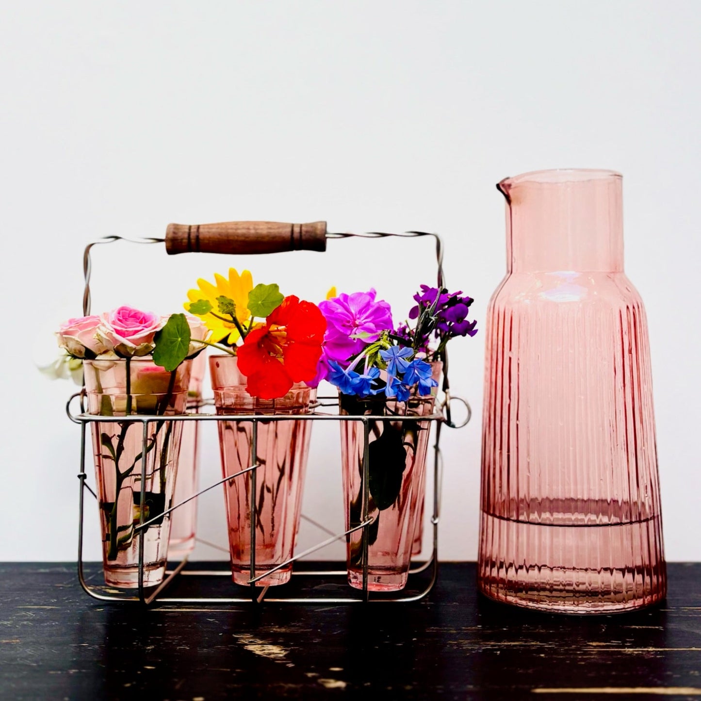 Pink glass carafe with a metal caddy holding colorful flowers on a wooden surface.