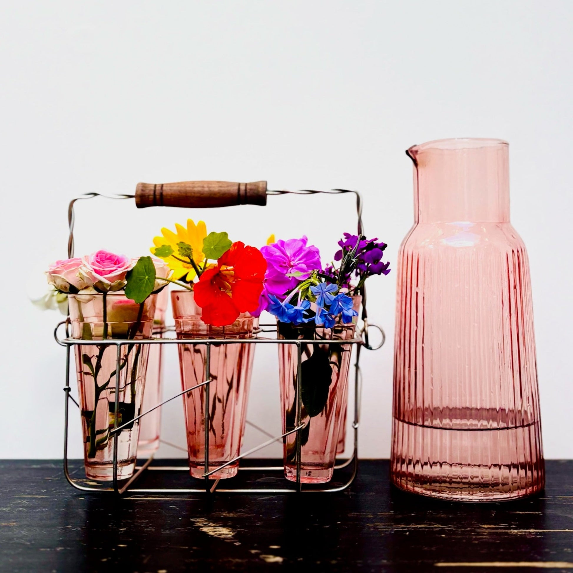 Pink glass carafe with a metal caddy holding colorful flowers on a wooden surface.