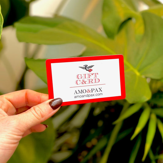 Hand holding a gift card with 'AMO & PAX' branding against a green leafy background