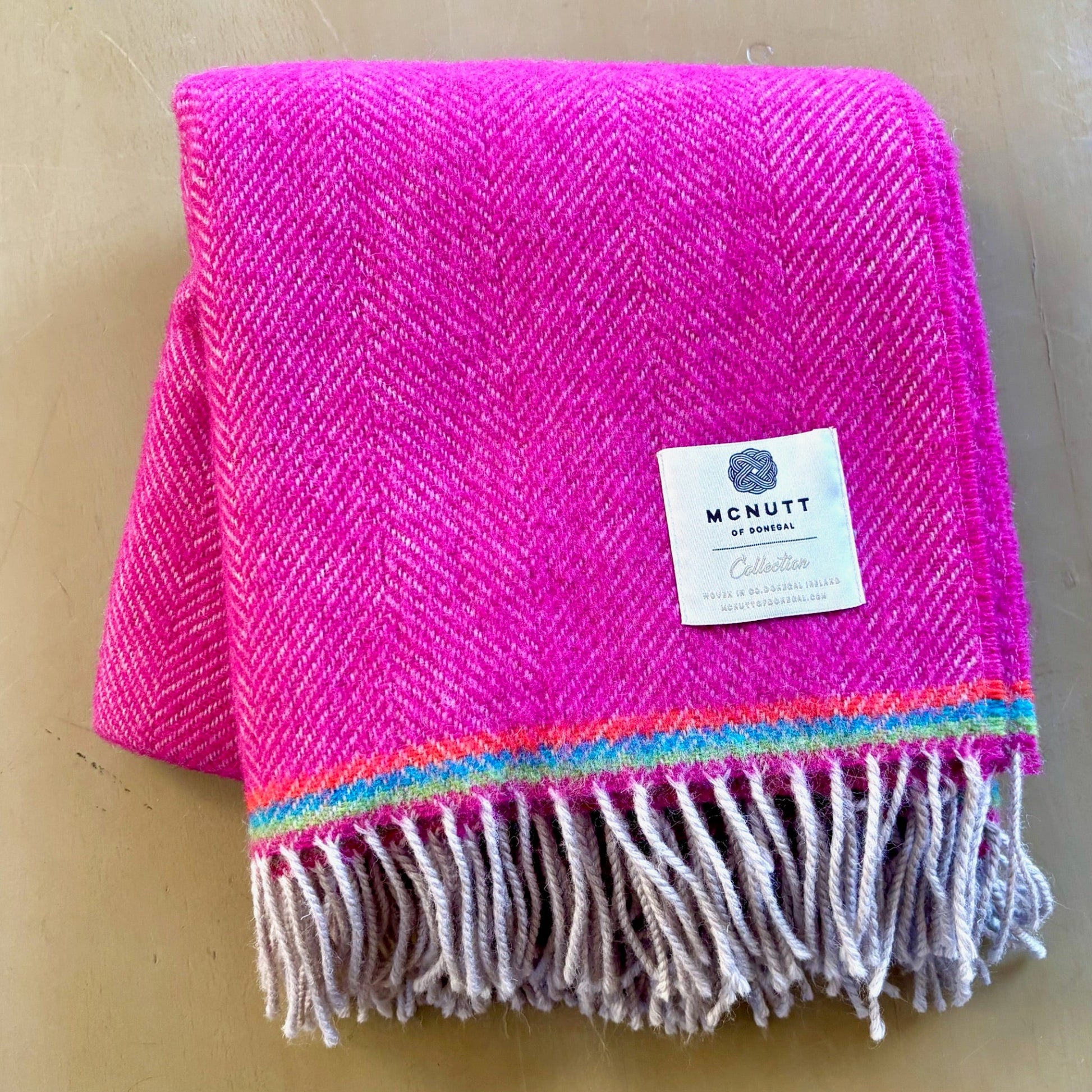 Pink woven blanket with colorful fringe and a visible brand label on a beige background