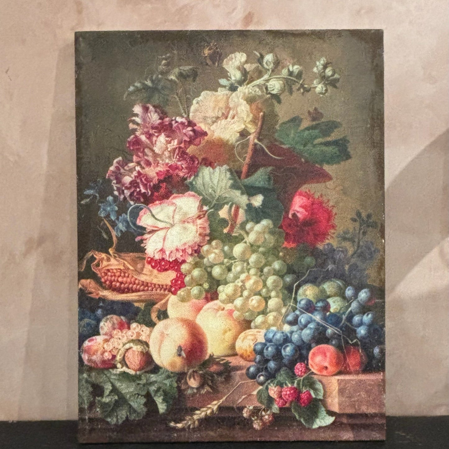 Framed still life painting of fruits and flowers on a textured wall.