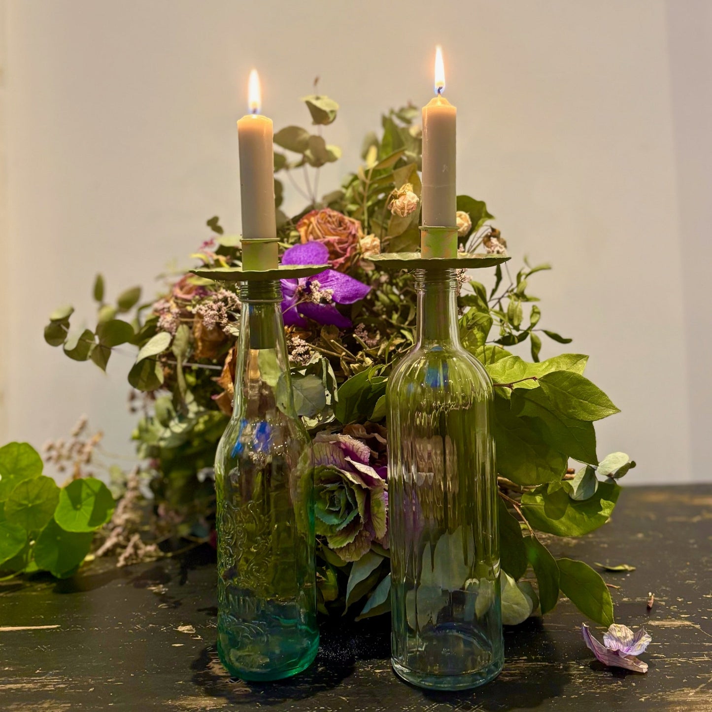 Two lit candles in green glass bottles with a floral arrangement in the background.