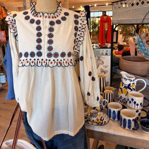 White blouse with floral embroidery on a mannequin, surrounded by ceramic items in a store setting.