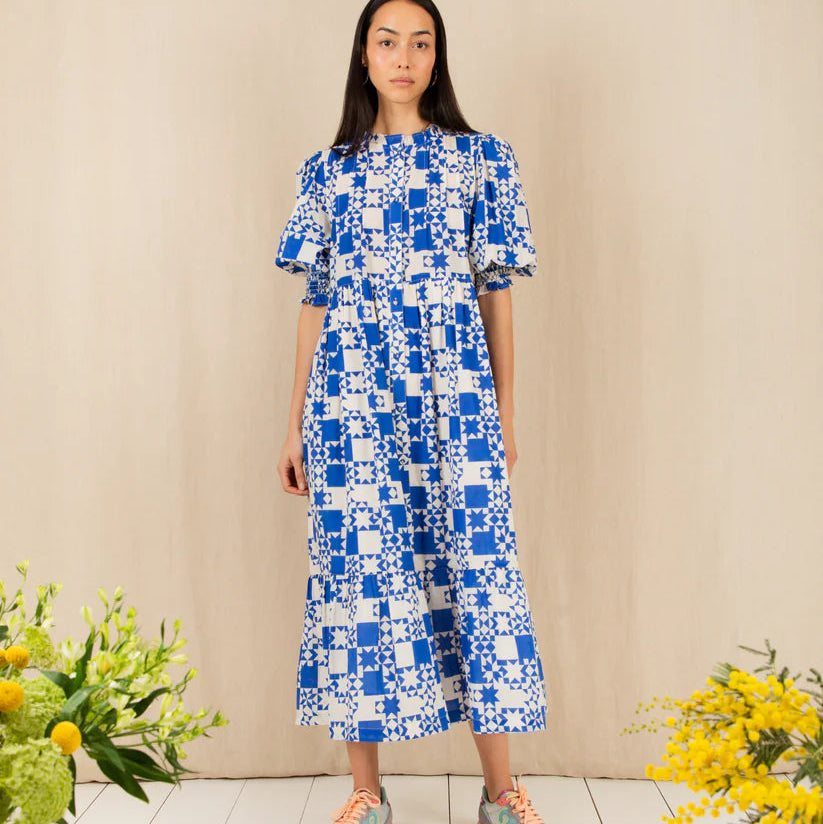 Woman wearing a blue and white patterned dress standing between two flower arrangements on a beige background.