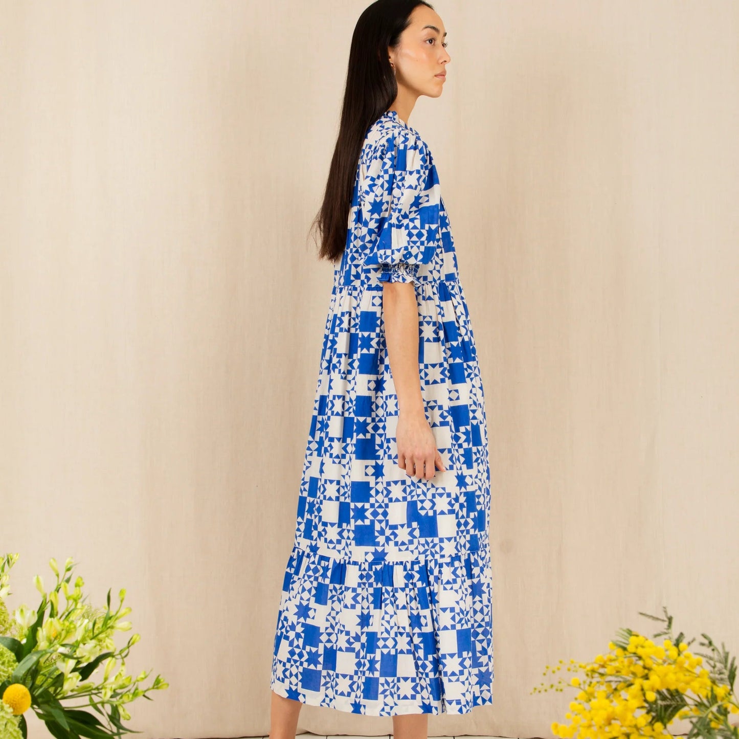 Woman wearing a blue and white patterned dress standing against a beige wall with yellow flowers on either side.