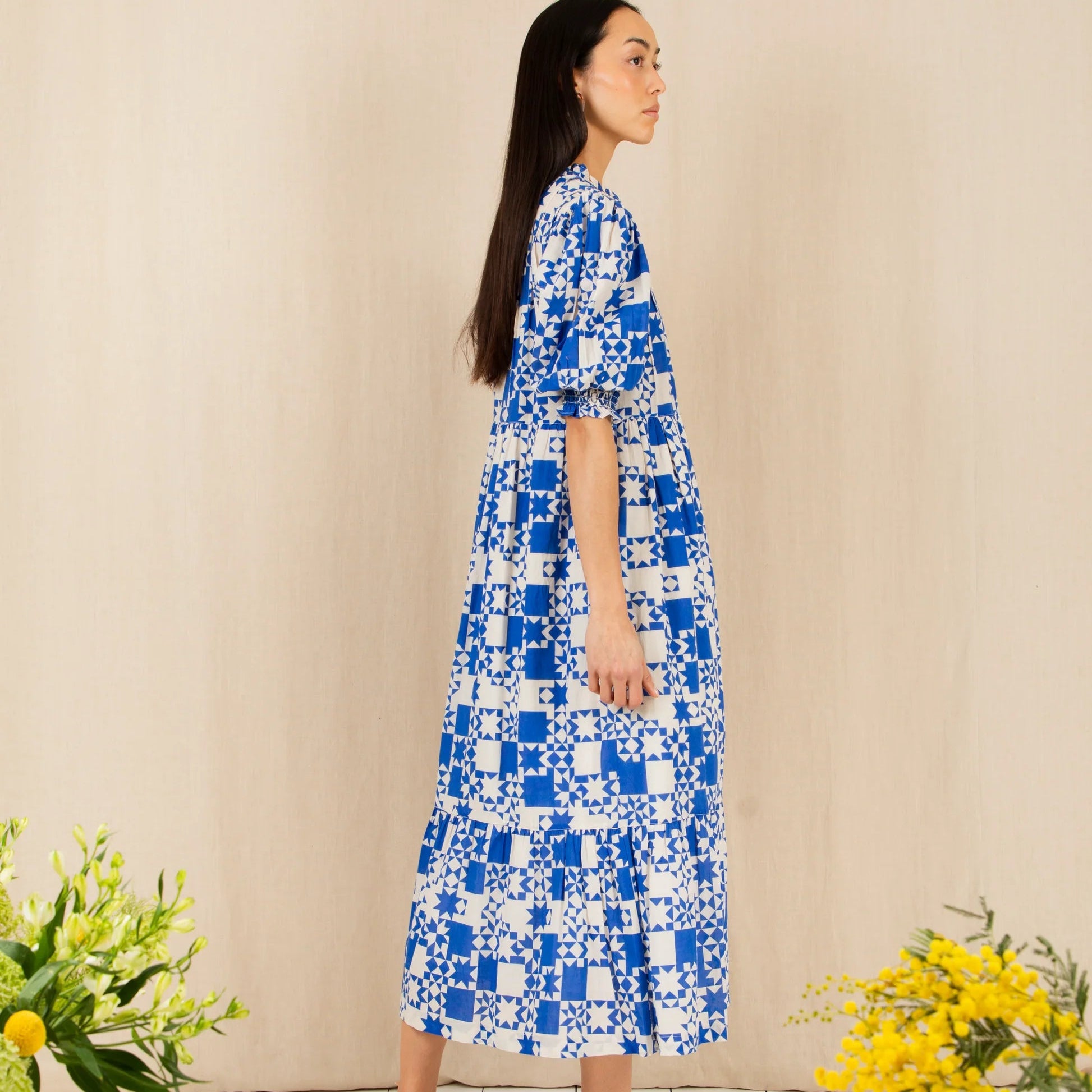 Woman wearing a blue and white patterned dress standing against a beige wall with yellow flowers on either side.
