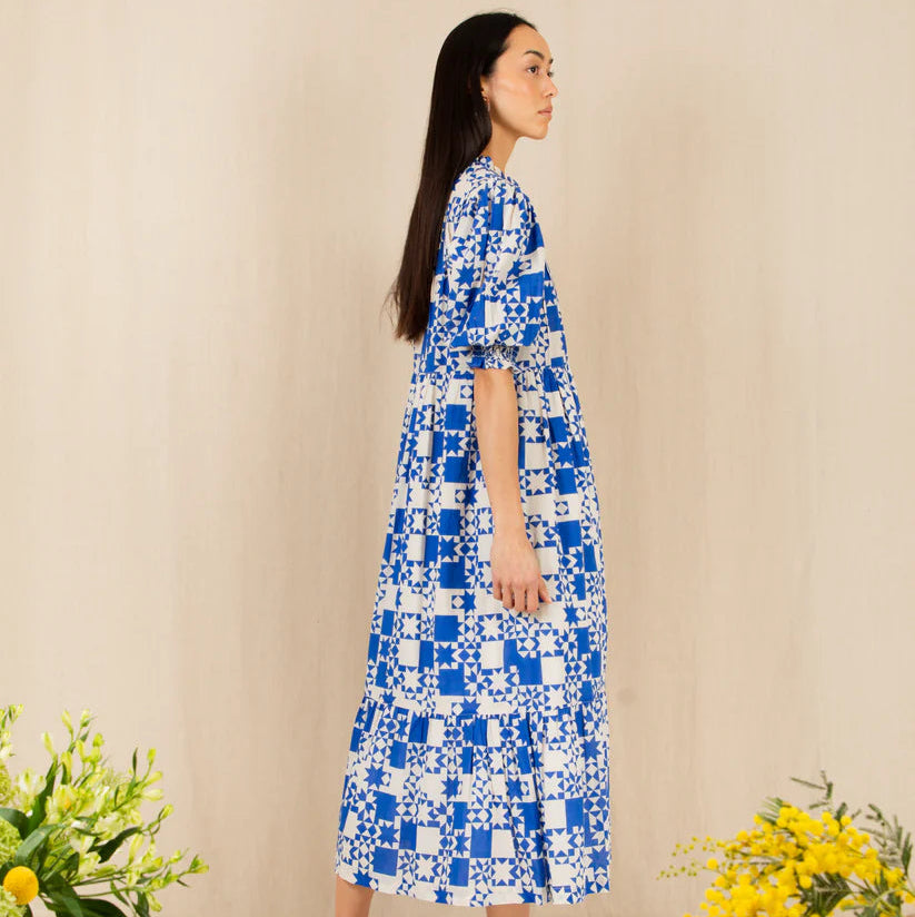 Woman wearing a blue and white patterned dress standing against a beige wall with yellow flowers on either side.