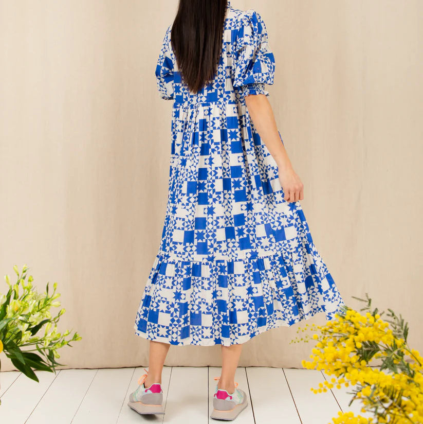 Woman wearing a blue and white checkered dress standing against a beige wall with flowers on either side.